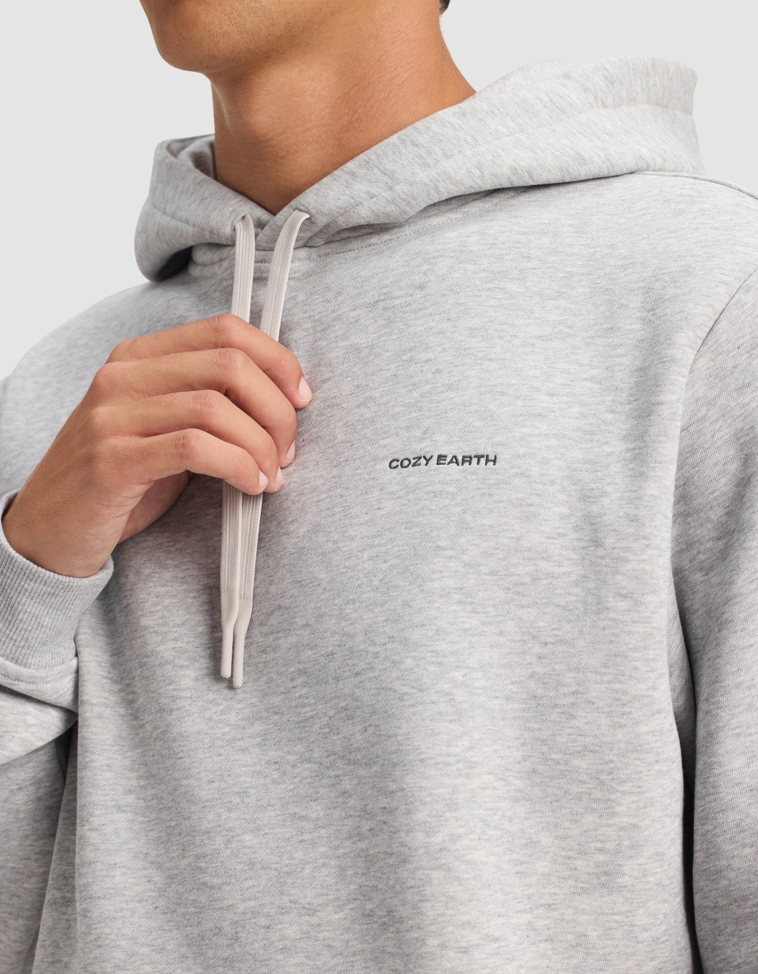 A person wearing the Cozy Earth Men's Cityscape 2.0 Hoodie in light gray holds the drawstrings near their neck against a plain, light-colored background. The "Cozy Earth" logo is visible on the chest.