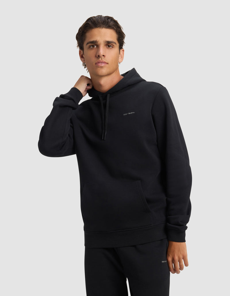 A young man models the Cozy Earth Men's Cityscape 2.0 Hoodie in black, paired with matching pants, standing against a light background with one hand on his neck and a neutral expression.