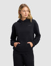 A young blonde woman models the Cozy Earth Women's Cityscape 2.0 Hoodie in black with matching pants, standing with hands in her hoodie pocket and looking slightly left, against a plain light grey background.|Color:Jet Black
