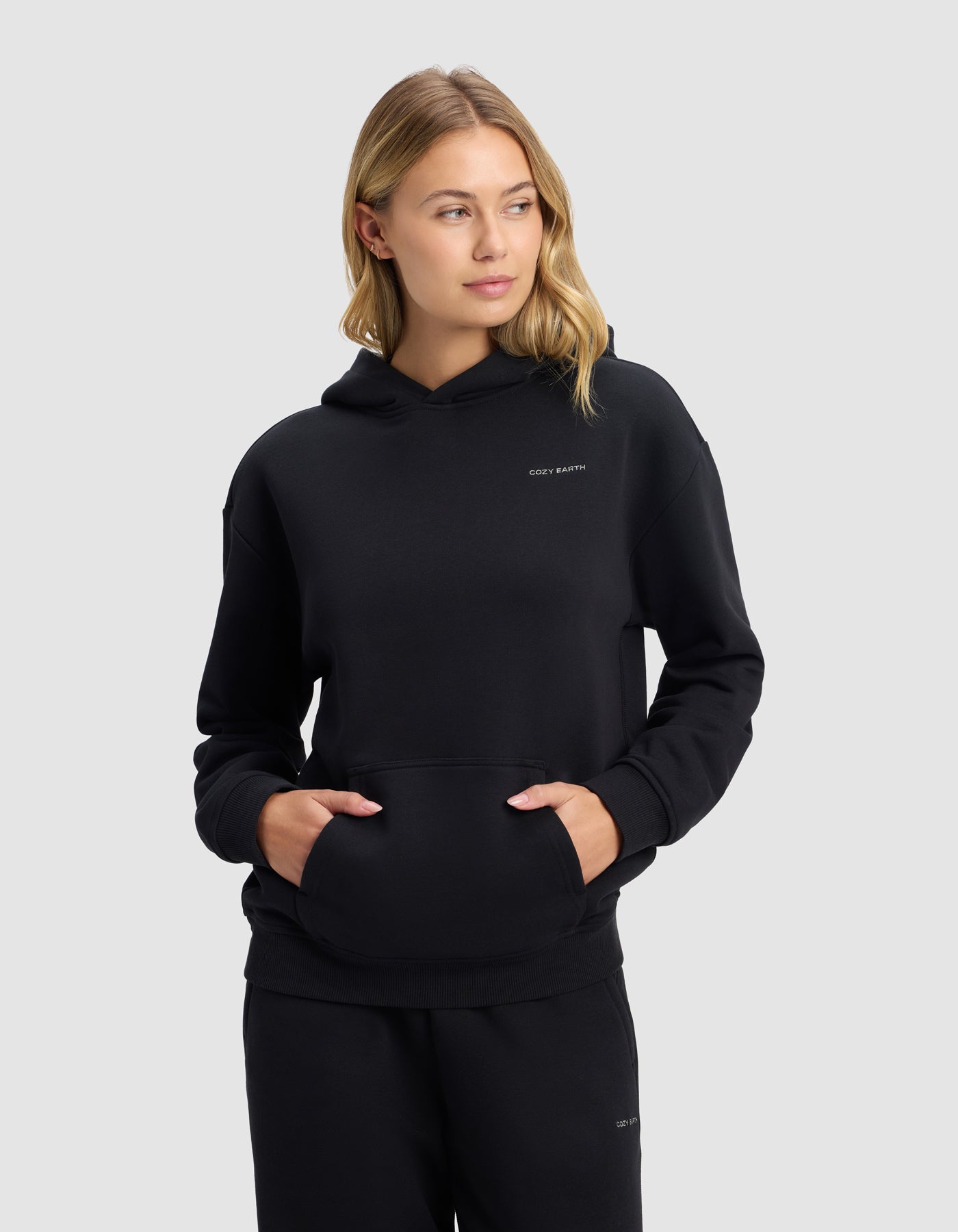 A young blonde woman models the Cozy Earth Women's Cityscape 2.0 Hoodie in black with matching pants, standing with hands in her hoodie pocket and looking slightly left, against a plain light grey background.|Color:Jet Black