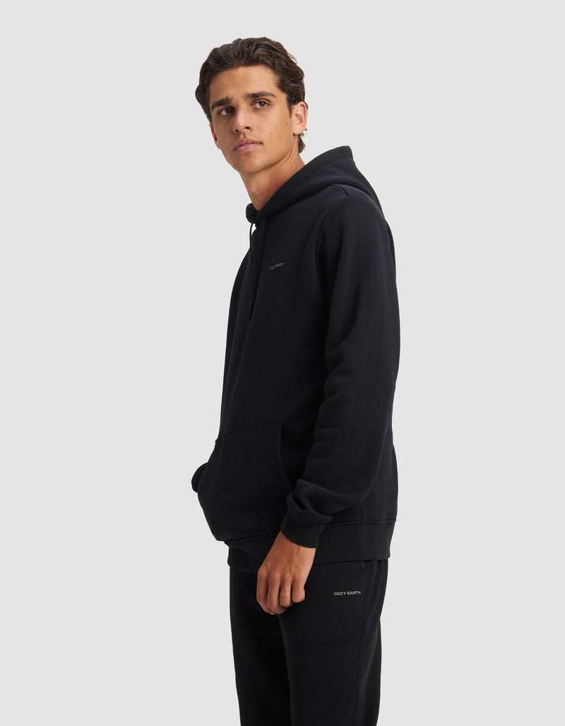 A young man with short brown hair wears the Cozy Earth Men's Cityscape 2.0 Hoodie and black pants, standing against a plain light gray background and looking slightly to his left with one hand in his hoodie pocket.