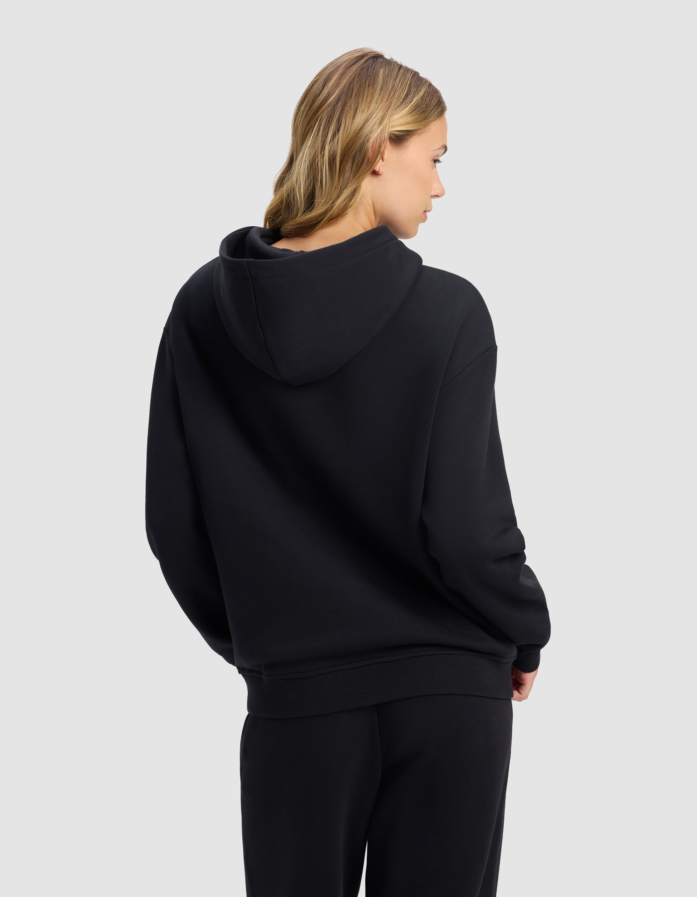 A woman with blonde hair models the Cozy Earth Women's Cityscape 2.0 Hoodie, standing in matching black pants with her back turned and looking to the side against a plain light gray background.|Color:Jet Black