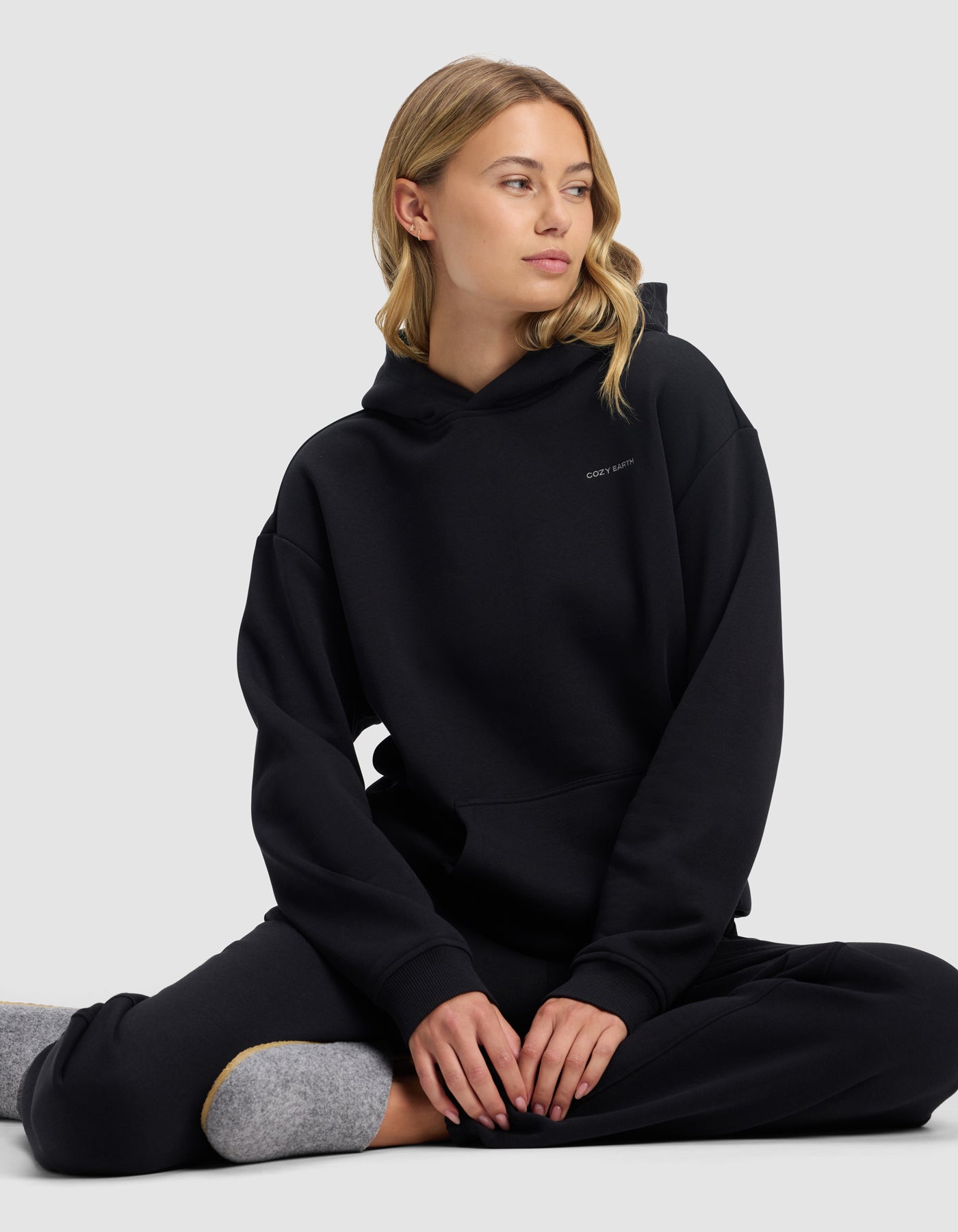 A woman with blonde hair sits cross-legged on the floor, wearing the Cozy Earth Women's Cityscape 2.0 Hoodie in black and matching pants. She gazes to the side with a neutral expression against a plain light gray background.|Color:Jet Black