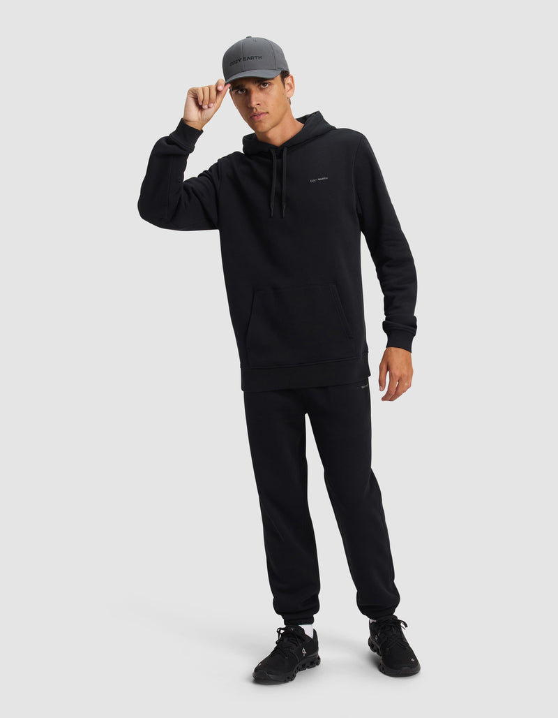A person wearing the Cozy Earth Men's Cityscape 2.0 Hoodie stands against a plain background, dressed in black joggers, black sneakers, and a gray cap, holding the brim of the cap with one hand.