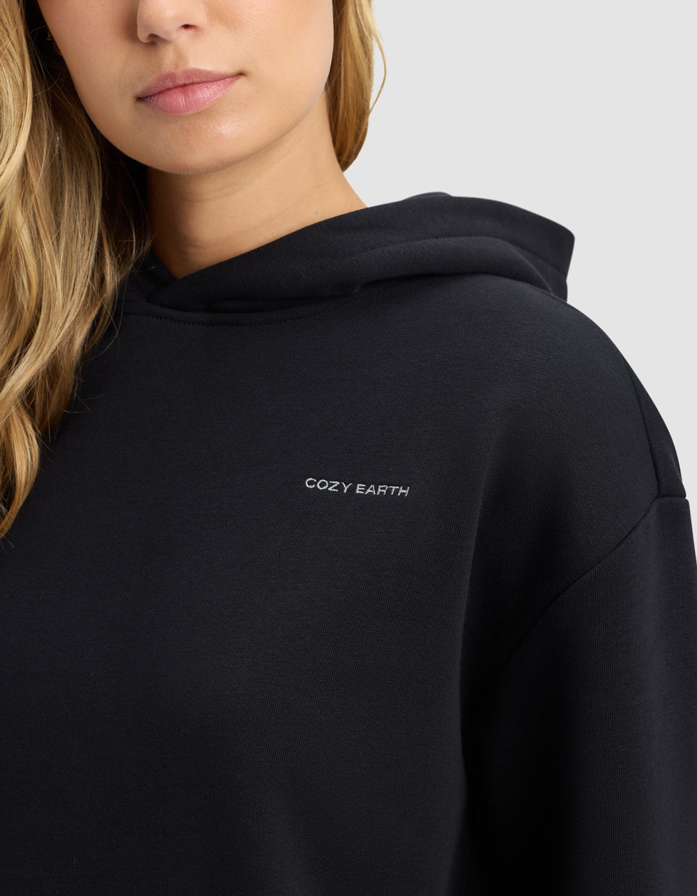 A woman with blonde hair wears the Cozy Earth Women's Cityscape 2.0 Hoodie in black, featuring small white "COZY EARTH" lettering on the chest. Only her lower face and upper body are visible against a light gray background.