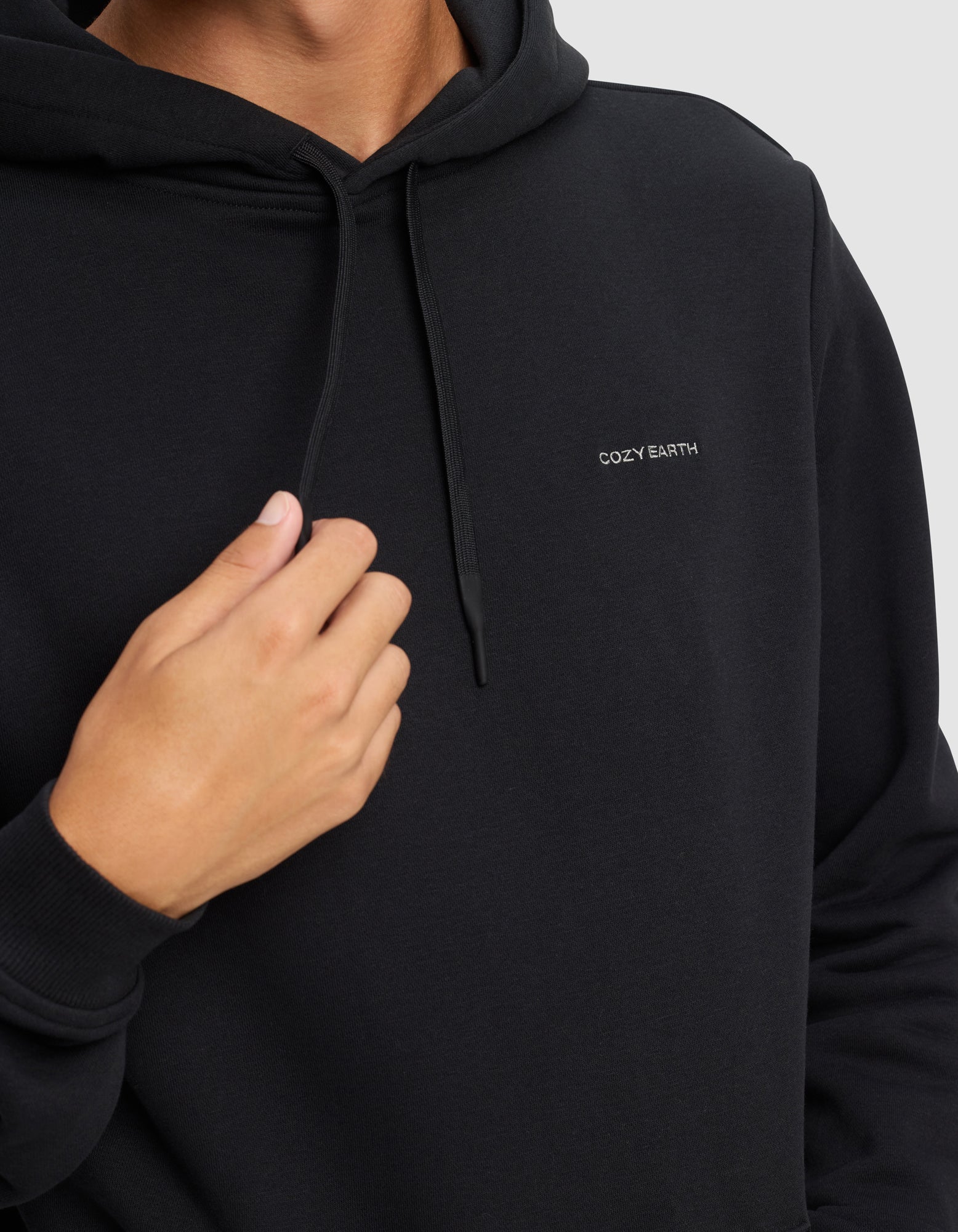 A man wears the Men's Cityscape 2.0 Hoodie by Cozy Earth, featuring "COZY EARTH" embroidered in white on the chest, holding the drawstring with one hand against a plain light background.