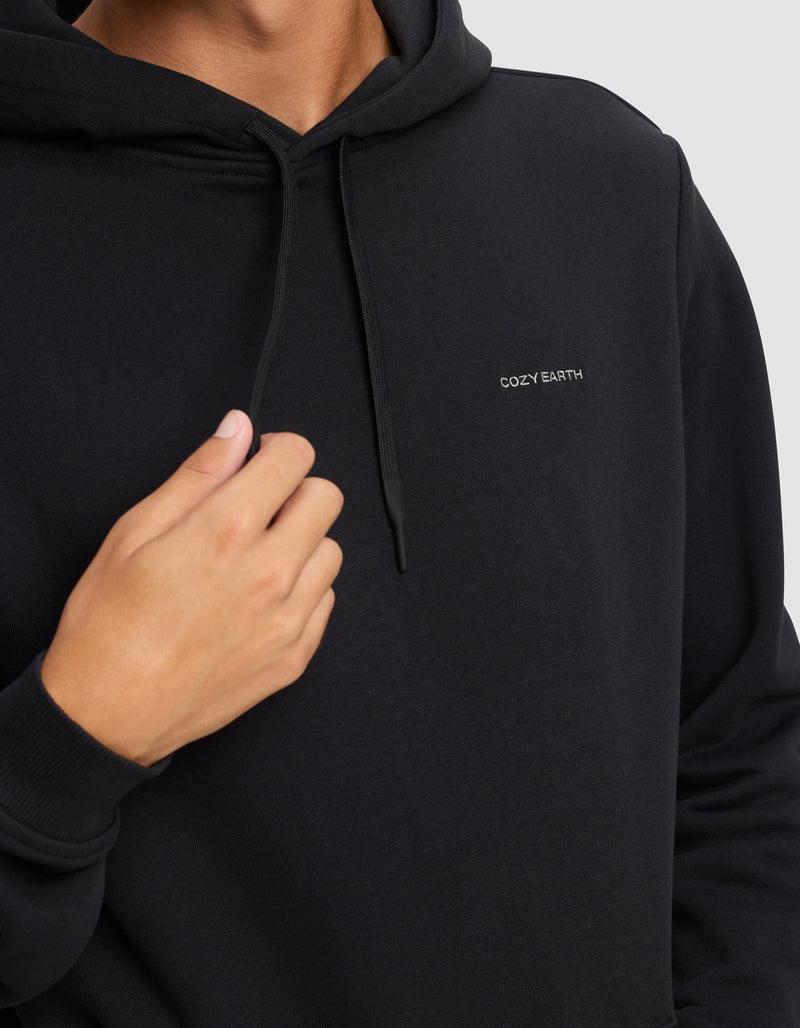 A man wears the Men's Cityscape 2.0 Hoodie by Cozy Earth, featuring "COZY EARTH" embroidered in white on the chest, holding the drawstring with one hand against a plain light background.
