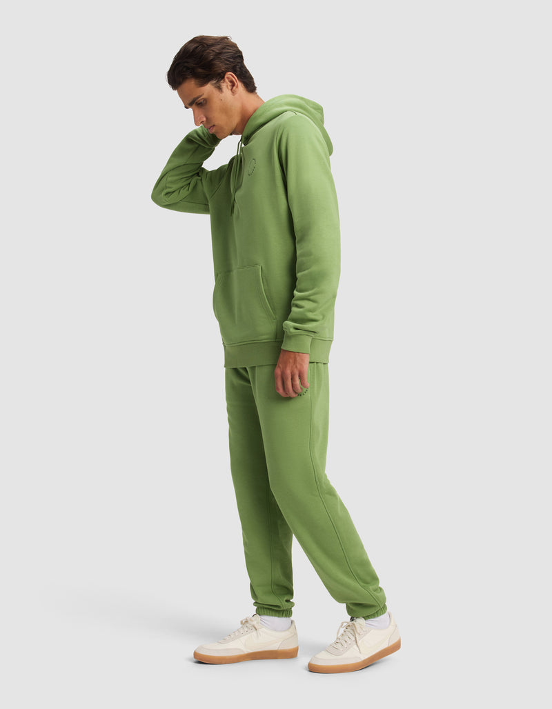 A man wears the Cozy Earth Men's Cityscape 2.0 Hoodie & Sweatpant Set in light green, paired with white sneakers with tan soles, standing against a plain light background and touching the back of his neck.