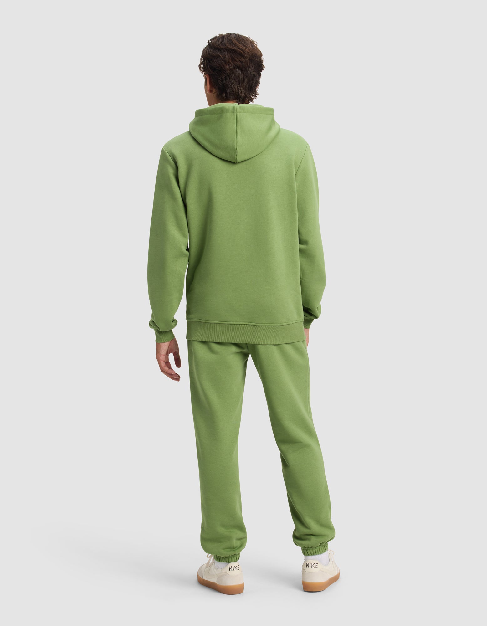 A person with short brown hair is standing and facing away, wearing Cozy Earth's Men's Cityscape 2.0 Hoodie & Sweatpant Set in light green and white sneakers with beige soles, against a plain light background.