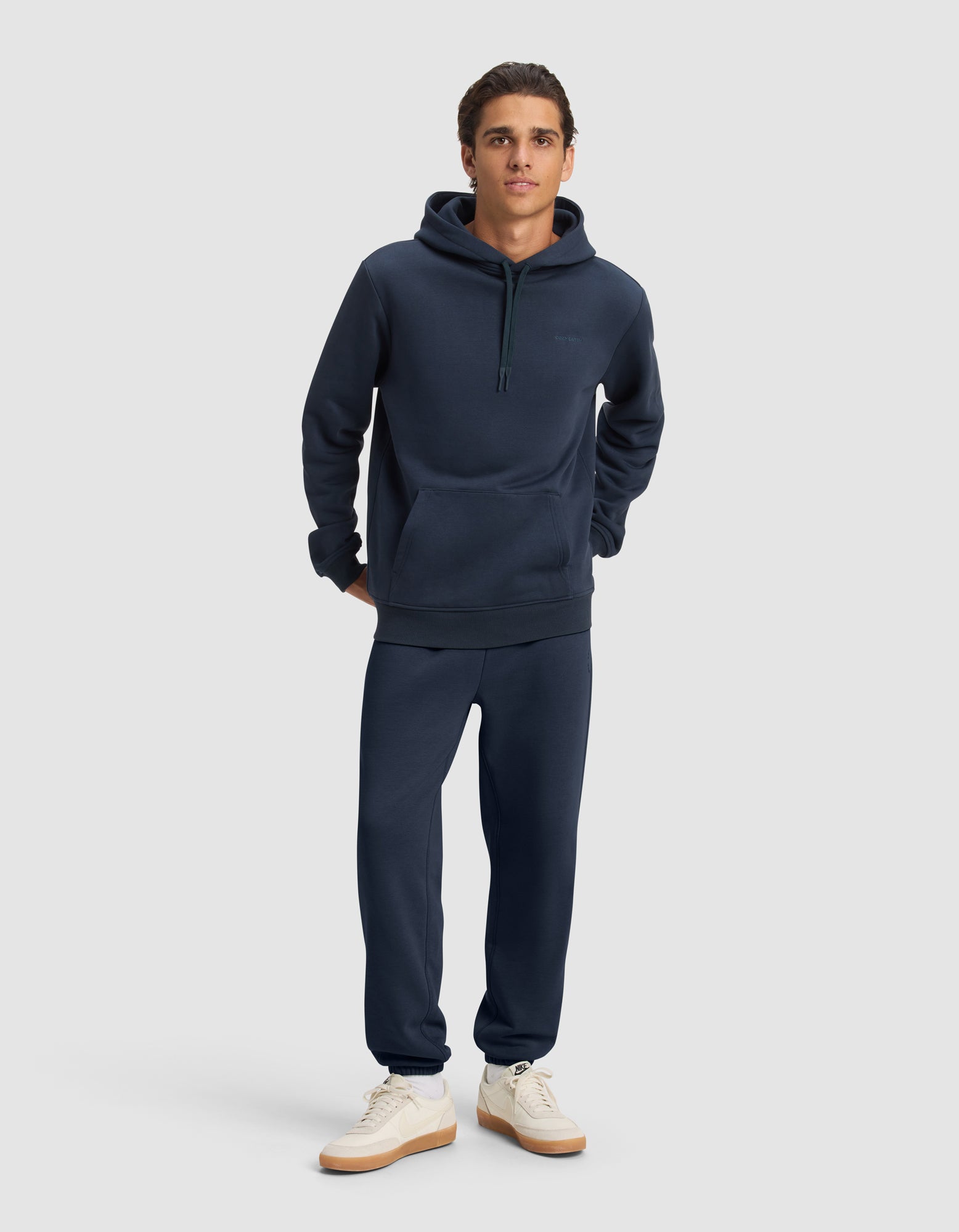 A young man poses against a light gray background, wearing the Cozy Earth Men's Cityscape 2.0 Hoodie & Sweatpant Set in navy blue with white sneakers. His hands are behind his back, and he faces forward.|Color:Eclipse