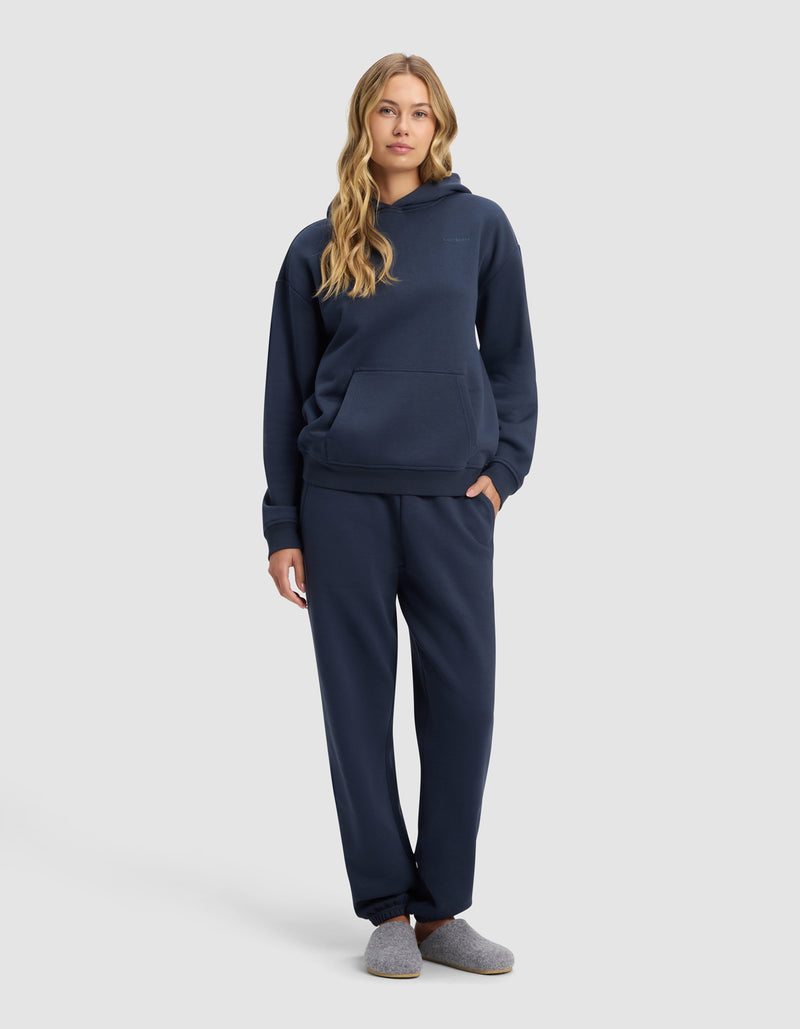 A woman with long wavy hair stands facing forward, wearing Cozy Earth’s Women’s Cityscape 2.0 Hoodie & Sweatpant Set in navy blue, paired with gray slippers, against a plain light gray background.