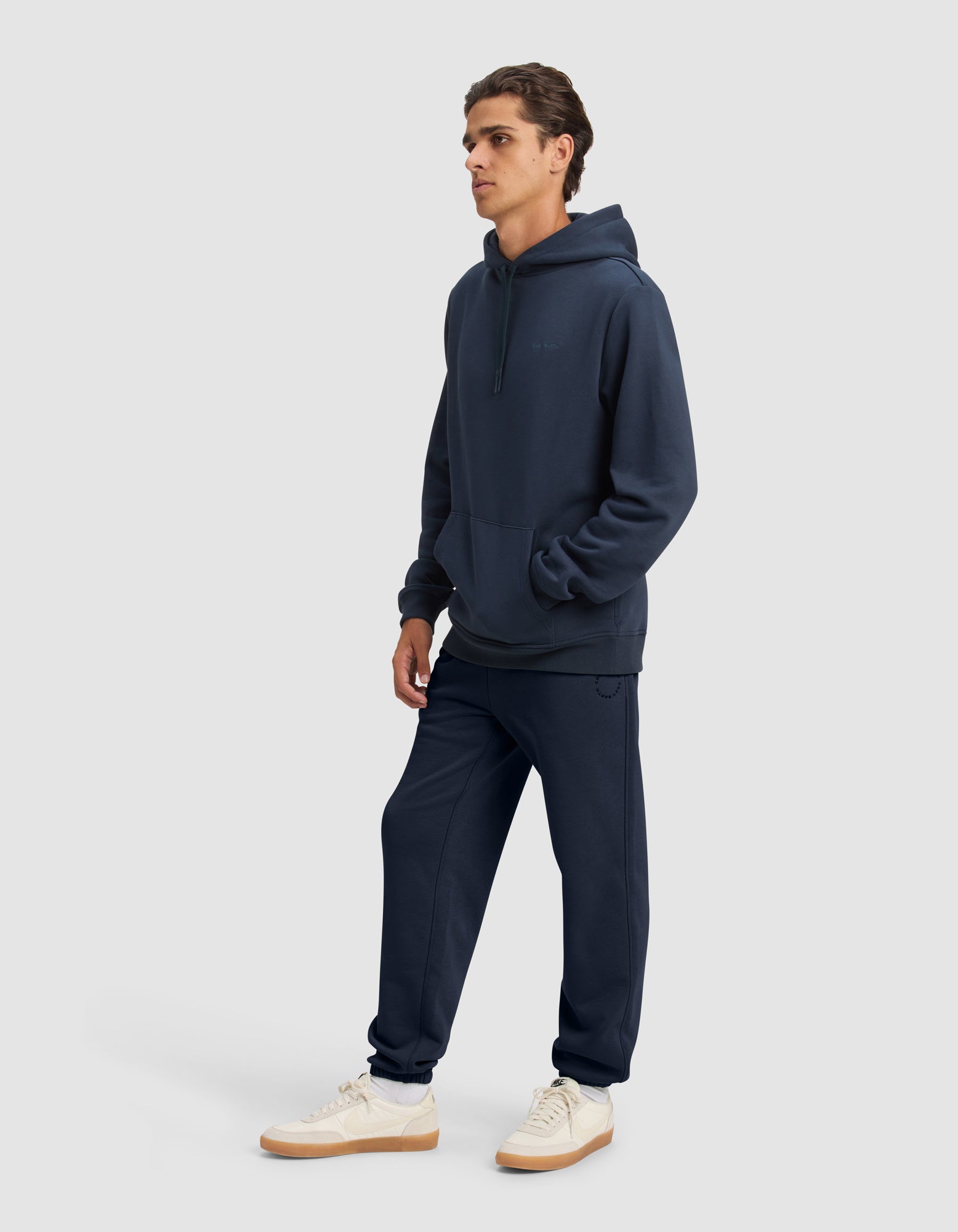 A man stands against a plain light gray background, wearing the Cozy Earth Men's Cityscape 2.0 Hoodie & Sweatpant Set in dark navy, paired with white sneakers featuring light brown soles, facing slightly to the side with hands in his pockets.