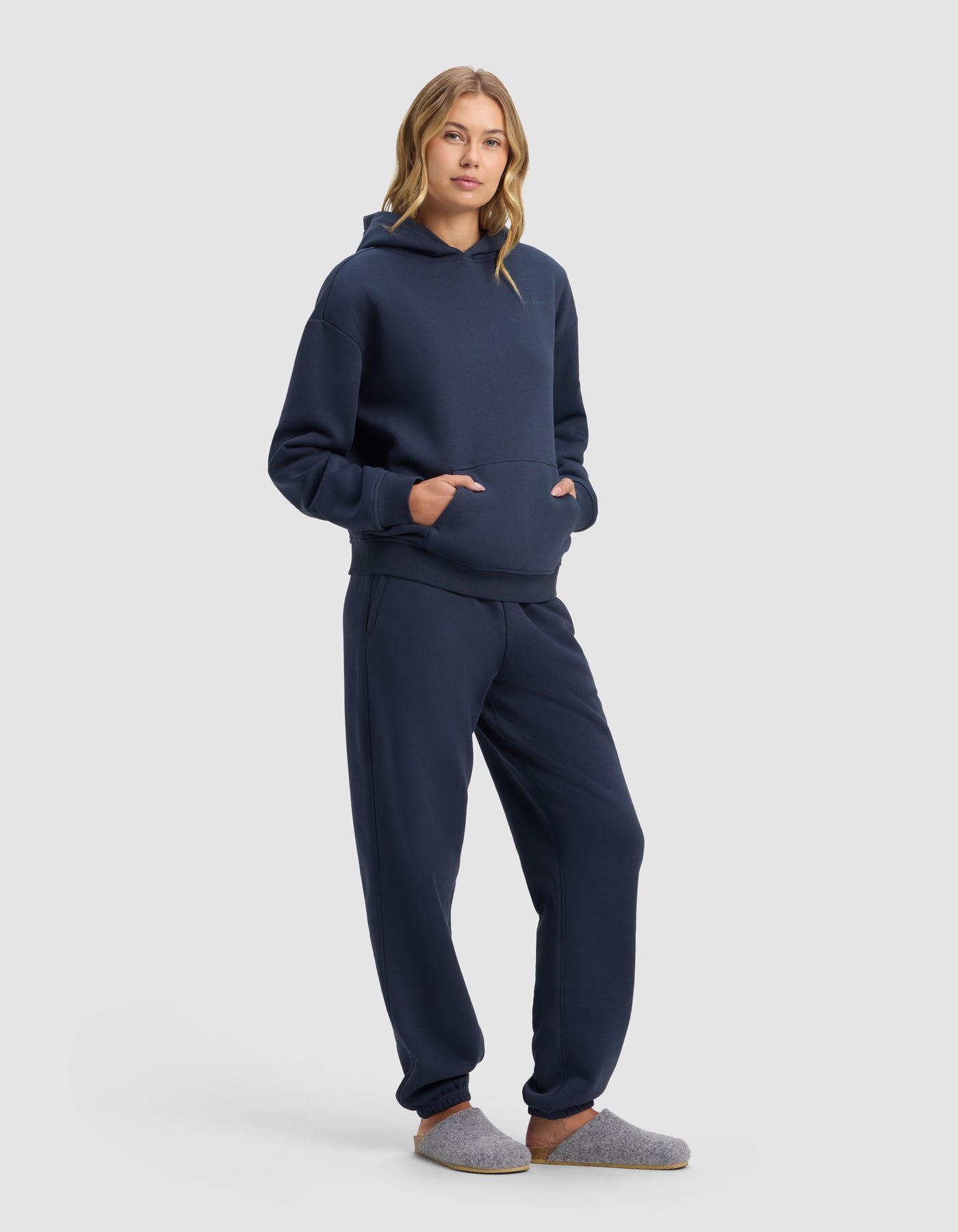 A person with long, wavy hair stands against a plain light background wearing Cozy Earth’s Women’s Cityscape 2.0 Hoodie & Sweatpant Set in navy blue, hands in the hoodie pocket, and gray slippers.