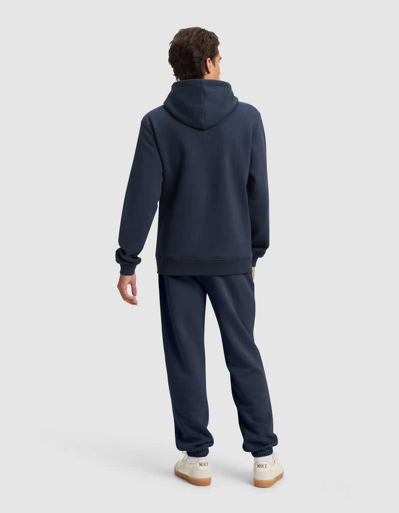A person faces away from the camera, wearing the Cozy Earth Men's Cityscape 2.0 Hoodie & Sweatpant Set in navy blue, paired with white Nike sneakers against a plain light gray background.