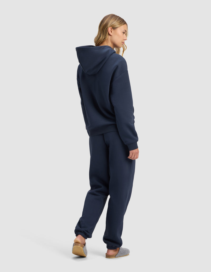 A person with long blonde hair faces away, wearing the Cozy Earth Women's Cityscape 2.0 Hoodie & Sweatpant Set in navy blue, styled with gray slippers against a plain light gray background.