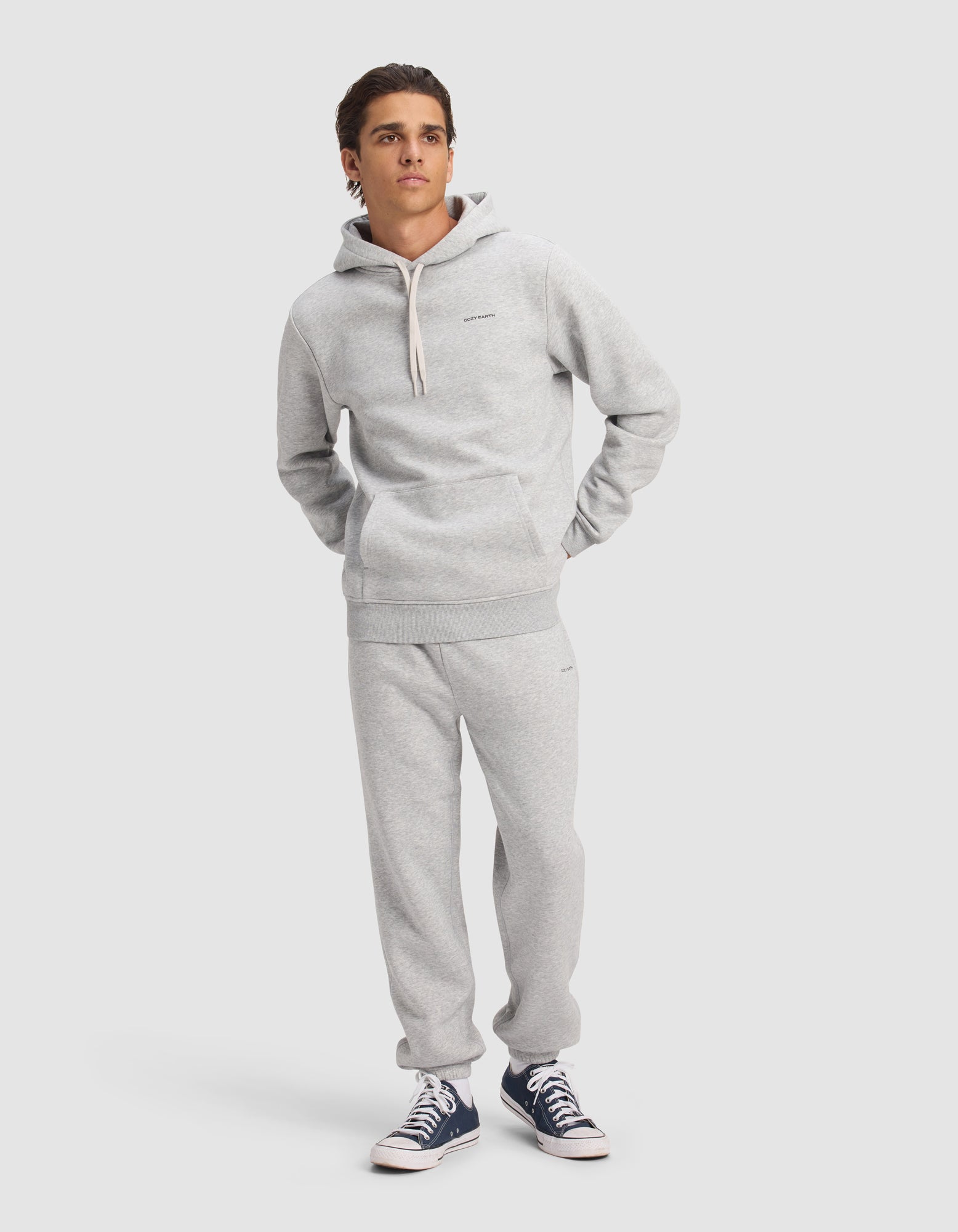 A young man models the Cozy Earth Men's Cityscape 2.0 Hoodie & Sweatpant Set in light gray, standing against a plain background with his hands in his pockets and navy blue sneakers, looking slightly to the side.