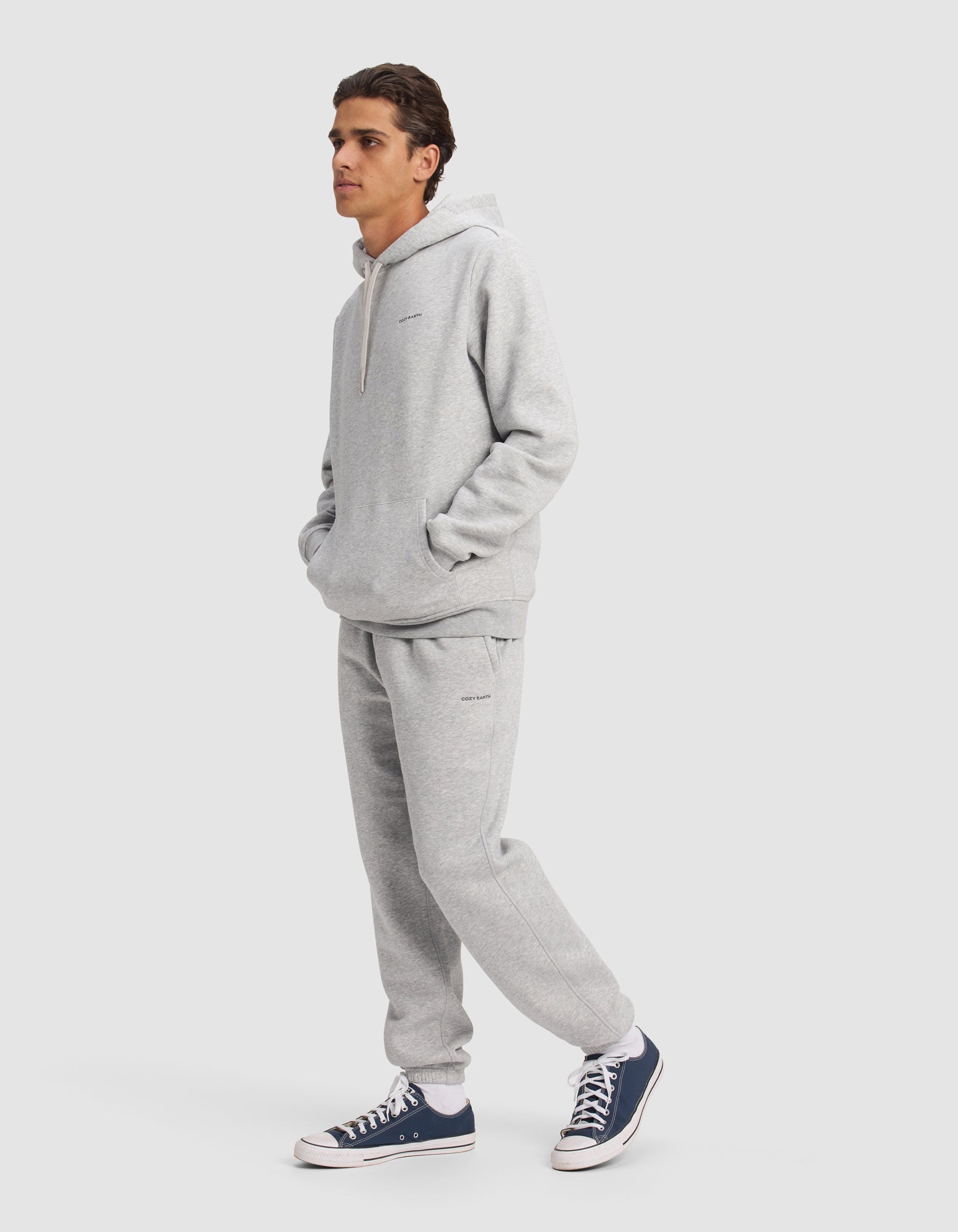 A man poses against a plain light background, hands in the pockets of a light gray Cozy Earth Men's Cityscape 2.0 Hoodie & Sweatpant Set, paired with navy blue and white sneakers.
