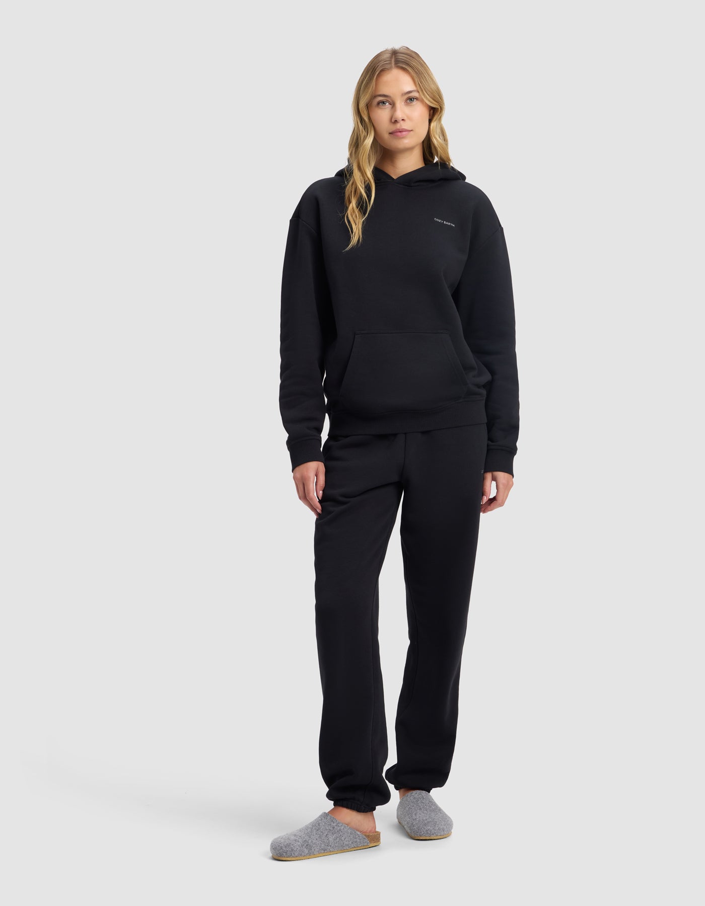 A woman with long blonde hair models the Cozy Earth Women's Cityscape 2.0 Hoodie & Sweatpant Set in black with grey slippers, standing against a plain light grey background.|Color:Jet Black