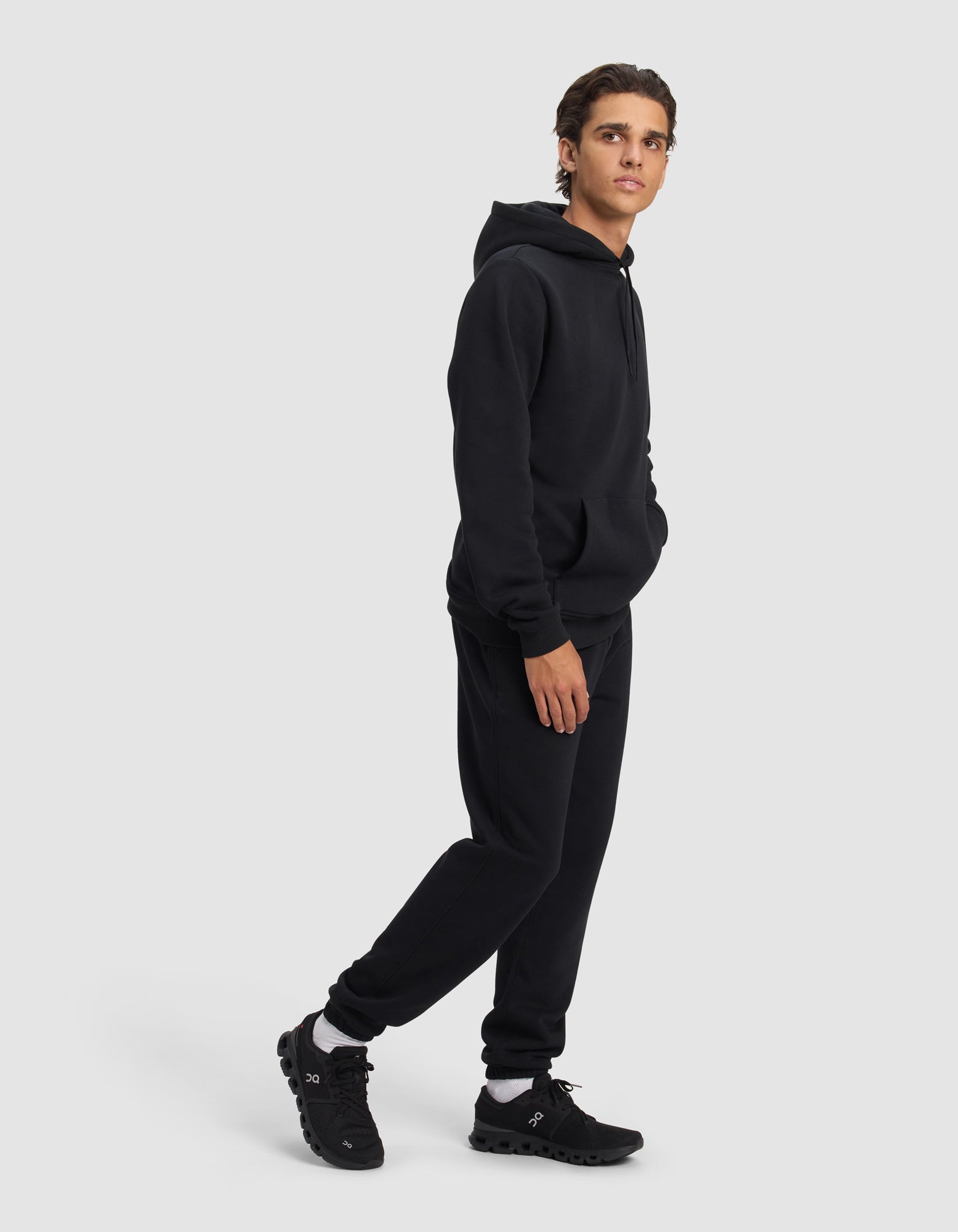 A young man models the Cozy Earth Men's Cityscape 2.0 Hoodie & Sweatpant Set in black, standing against a plain light gray background with one hand in his pocket, looking slightly to the side.