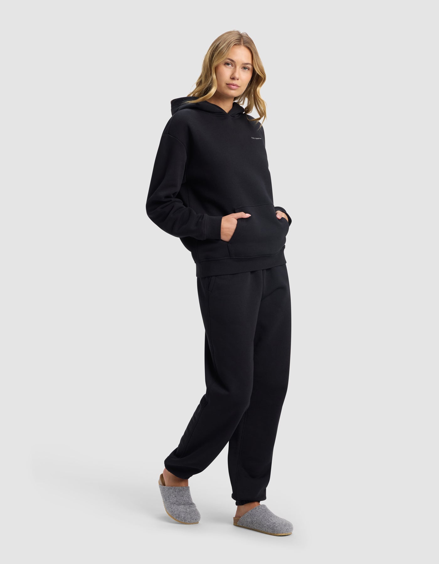 A woman with light brown hair poses against a light gray background wearing Cozy Earth's Women's Cityscape 2.0 Hoodie & Sweatpant Set in black, with her hands in the front hoodie pocket and gray slippers on her feet.