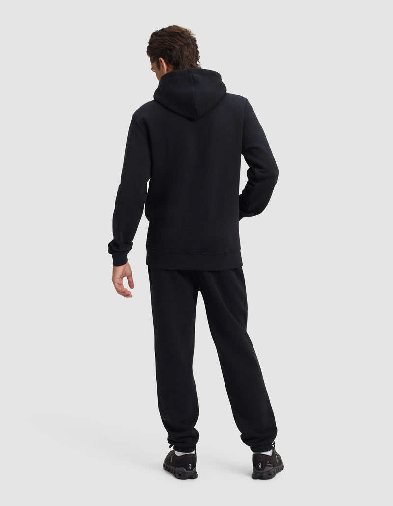 A person faces away from the camera, wearing Cozy Earth's Men's Cityscape 2.0 Hoodie & Sweatpant Set in black, with matching sneakers against a plain light gray background.