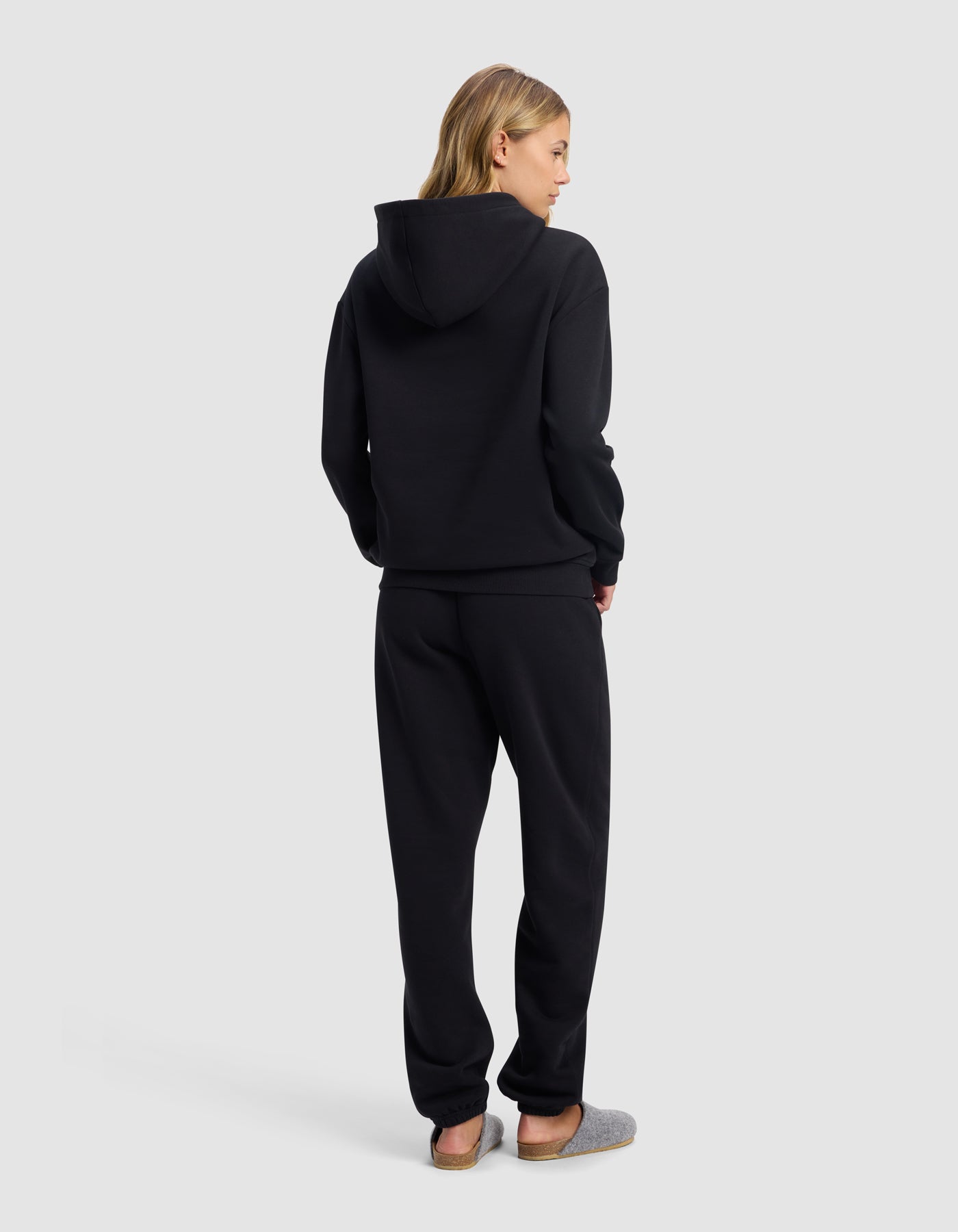 A woman with blonde hair, seen from behind, wears the Cozy Earth Women's Cityscape 2.0 Hoodie & Sweatpant Set in black with gray slippers, standing against a plain light gray background.