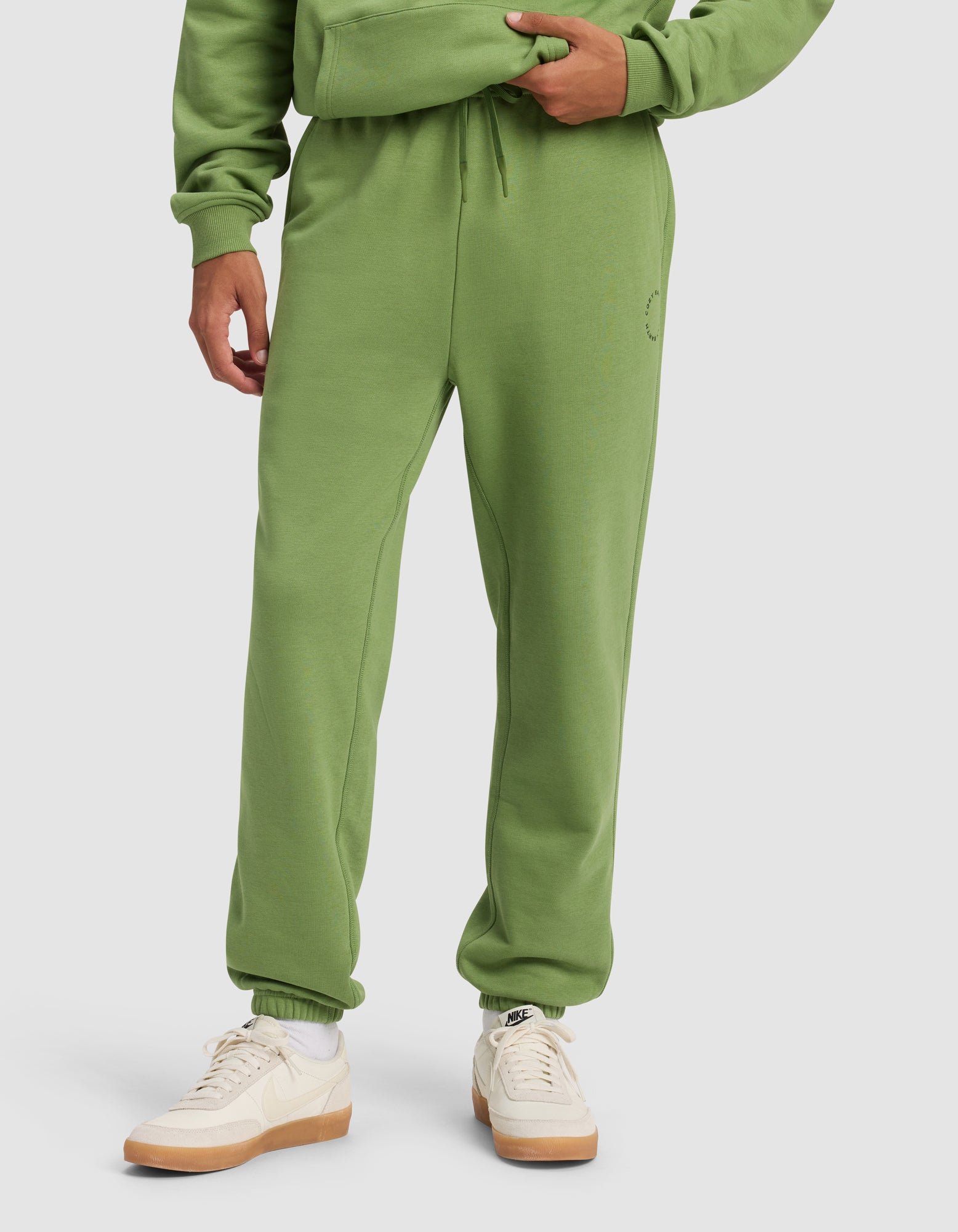 A person wears Cozy Earth's Men's Cityscape 2.0 Sweatpant in green, paired with a matching hoodie and white-beige Nike sneakers, standing against a plain light background. Only the lower half of their body is visible.