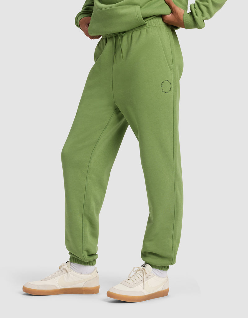 A person wearing Cozy Earth Men's Cityscape 2.0 Sweatpant in green with a matching sweatshirt, white socks, and beige sneakers stands hands on hips against a plain light background.