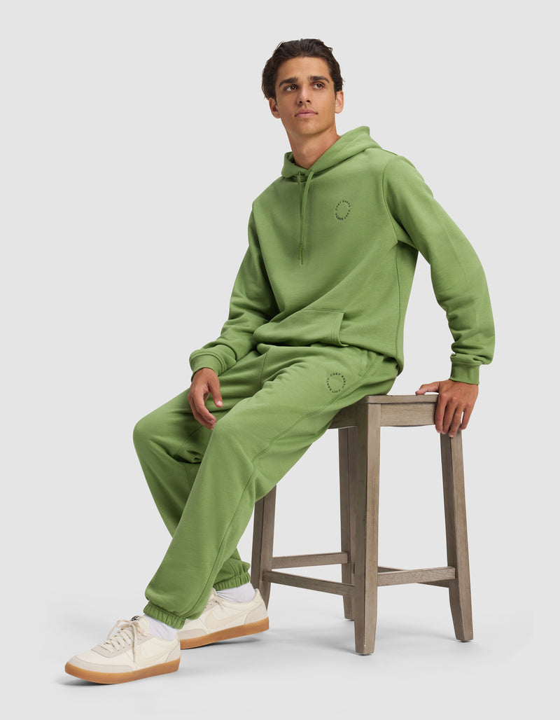 A person in Cozy Earth's Men's Cityscape 2.0 Sweatpant and a matching light green hoodie sits on a wooden stool, wearing white sneakers with beige soles. They have short brown hair, and the background is plain, light gray.
