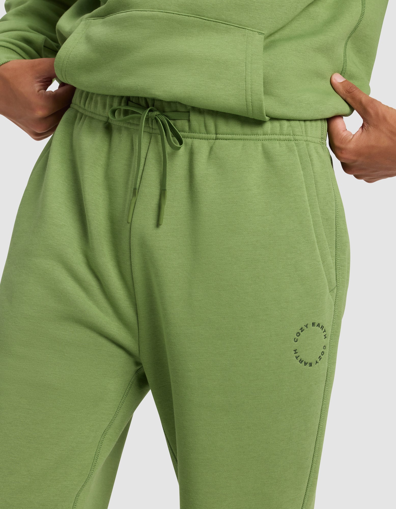 A person wears Cozy Earth Men's Cityscape 2.0 Sweatpant in light green with an elastic drawstring waistband, a small circular logo by the left pocket, and a matching sweatshirt, one hand in the pocket.