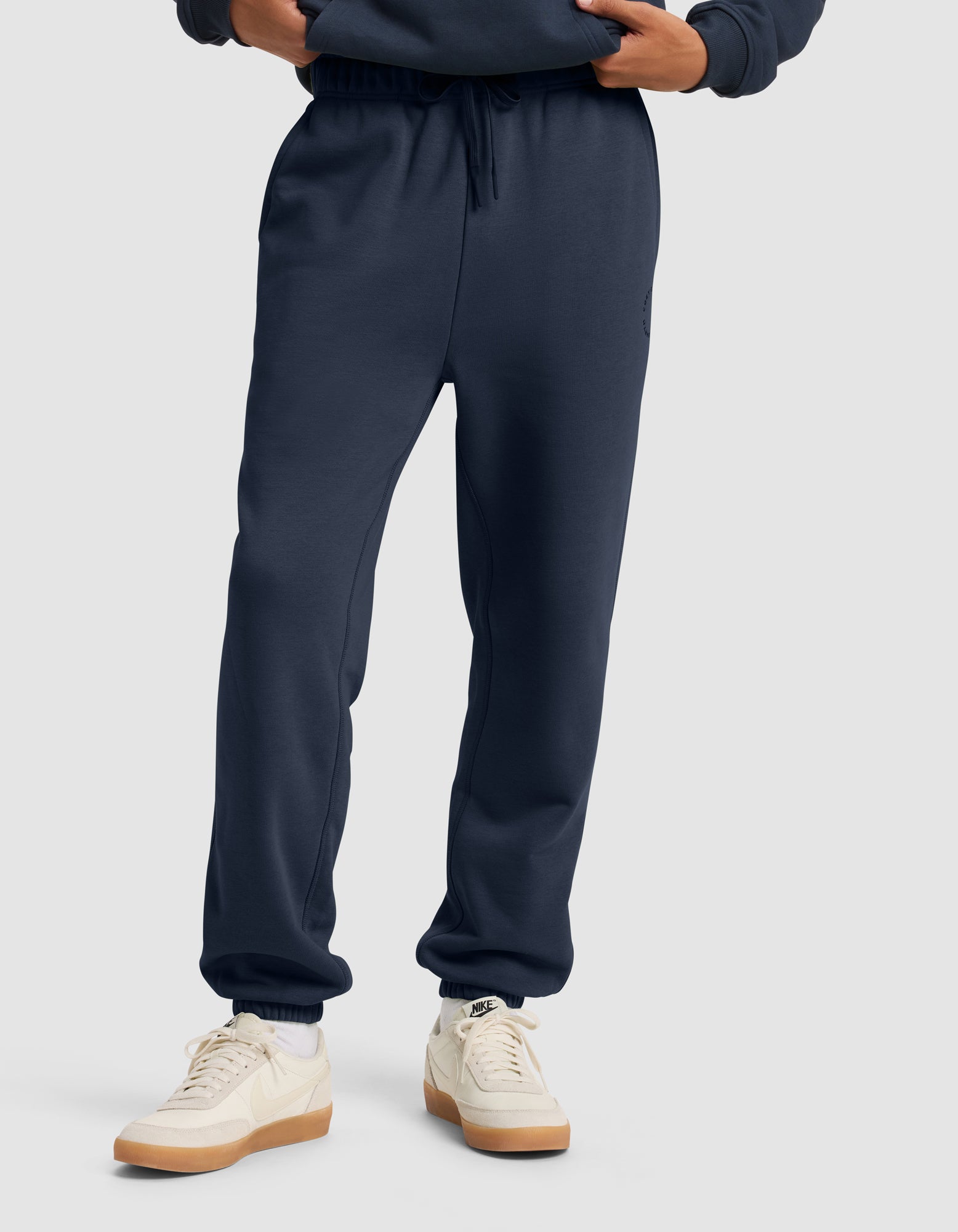 A man models Cozy Earth’s Men's Cityscape 2.0 Sweatpant in dark blue with white sneakers, hands in pockets, against a plain light gray background. Only his lower body is fully visible.|Color:Eclipse