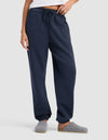 A woman stands against a plain light background wearing Cozy Earth’s Women’s Cityscape 2.0 Sweatpant in navy blue with an elastic waistband and drawstring, paired with a white top and grey slippers.|Color:Eclipse