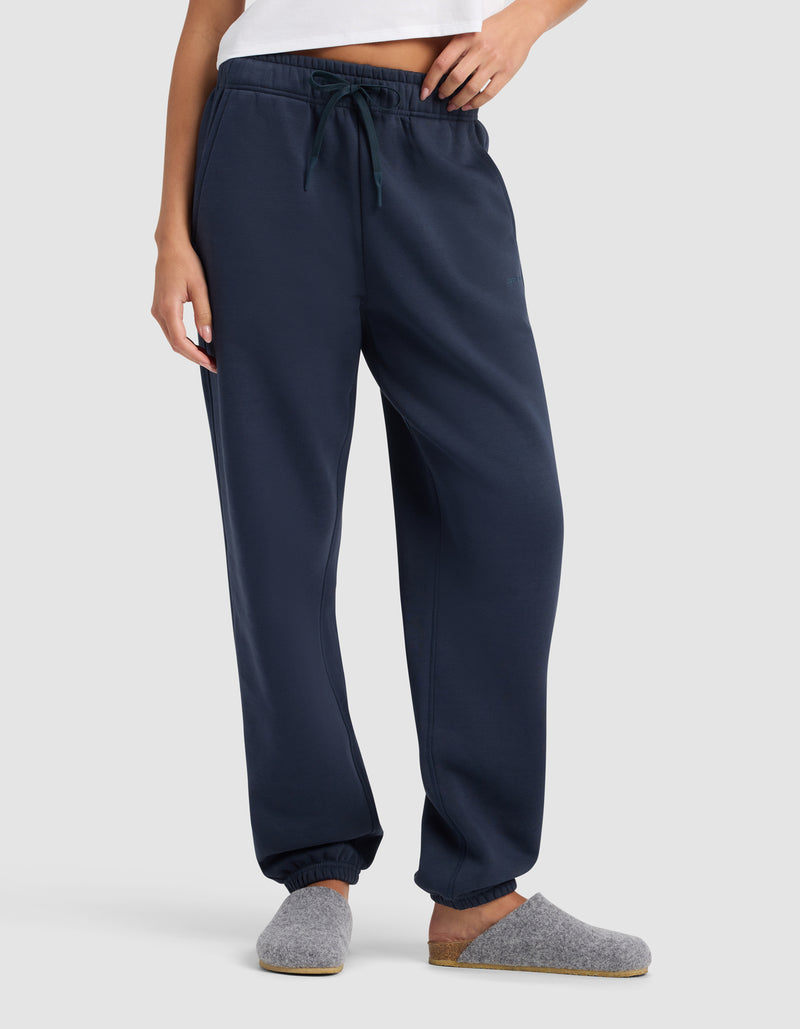 A woman stands against a plain light background wearing Cozy Earth’s Women’s Cityscape 2.0 Sweatpant in navy blue with an elastic waistband and drawstring, paired with a white top and grey slippers.