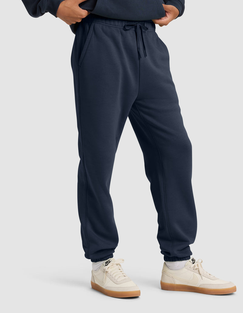 A man wears Cozy Earth's Men's Cityscape 2.0 Sweatpant in navy blue with a matching sweatshirt, white socks, and white sneakers with beige soles, standing against a plain light gray background.
