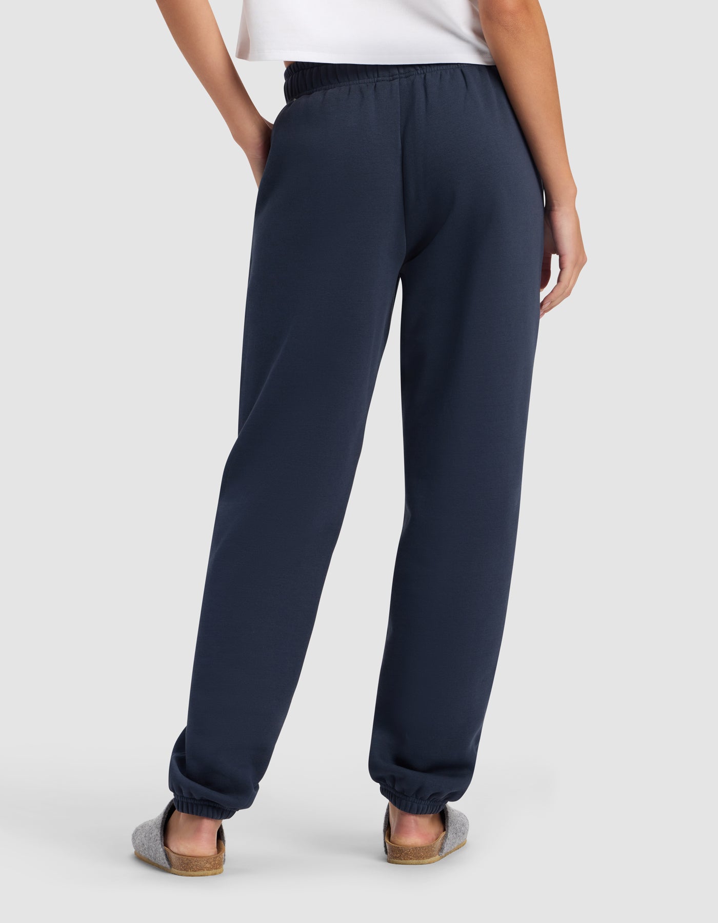 A person stands with their back to the camera, wearing a white shirt and Cozy Earth Women's Cityscape 2.0 Sweatpant in navy blue, paired with gray slippers against a light background.