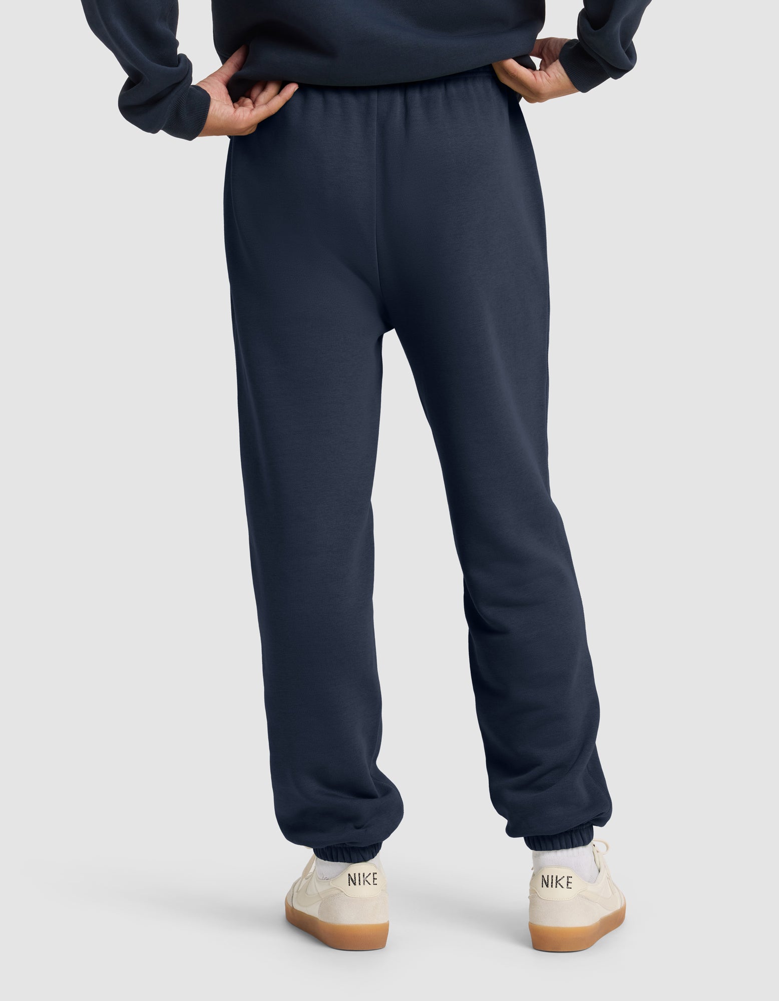 A person stands with hands on hips, wearing Cozy Earth Men's Cityscape 2.0 Sweatpant in navy and white Nike sneakers with tan soles, shown from the back against a plain light gray background.