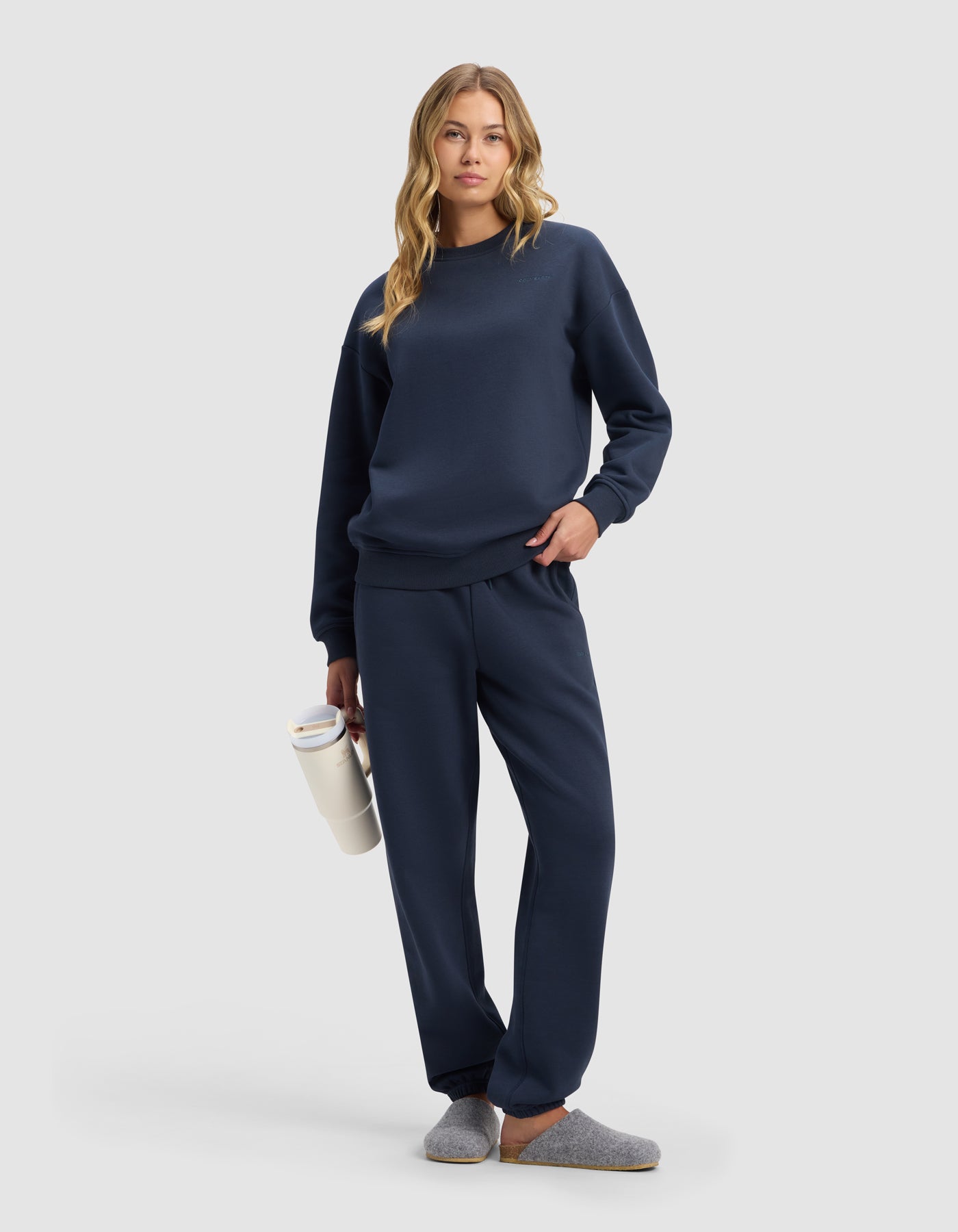 A woman in Cozy Earth's Women's Cityscape 2.0 Sweatpant, paired with a navy blue sweatshirt and gray slippers, holds a white water bottle. She has long wavy blonde hair, a relaxed look, and stands against a plain light gray background.
