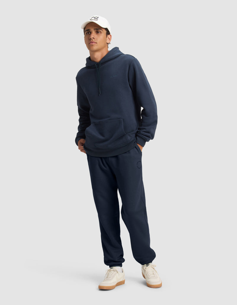 A man models the Cozy Earth Men's Cityscape 2.0 Sweatpant in navy blue with a matching hoodie, white baseball cap, and sneakers, standing against a plain gray background.