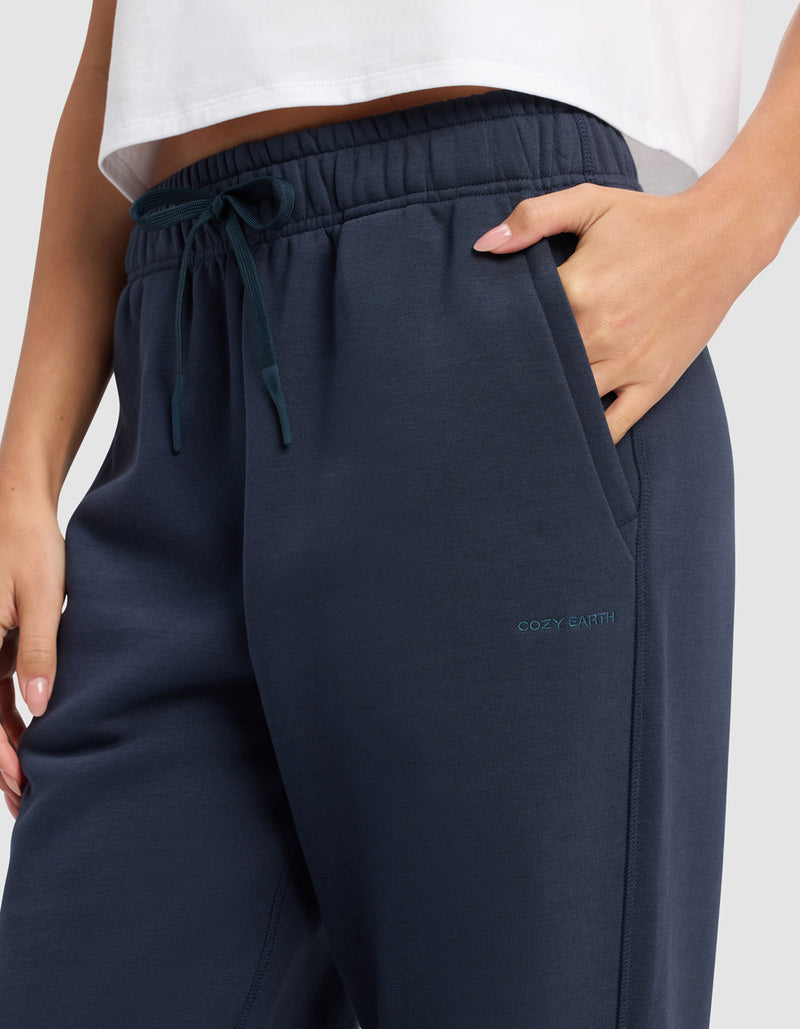 A person wears Cozy Earth Women's Cityscape 2.0 Sweatpant in dark blue with one hand in the pocket, visible drawstrings, part of a white top shown, and "COZY EARTH" embroidered near the pocket.
