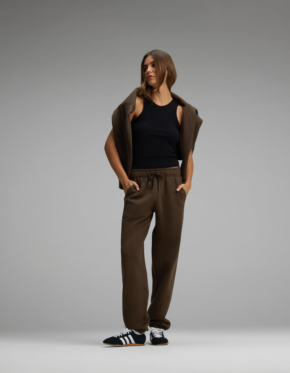 A person wears brown Cozy Earth Women’s CityScape Sweatpants with a drawstring waist, a black tank top, and a brown sweatshirt draped over their shoulders.