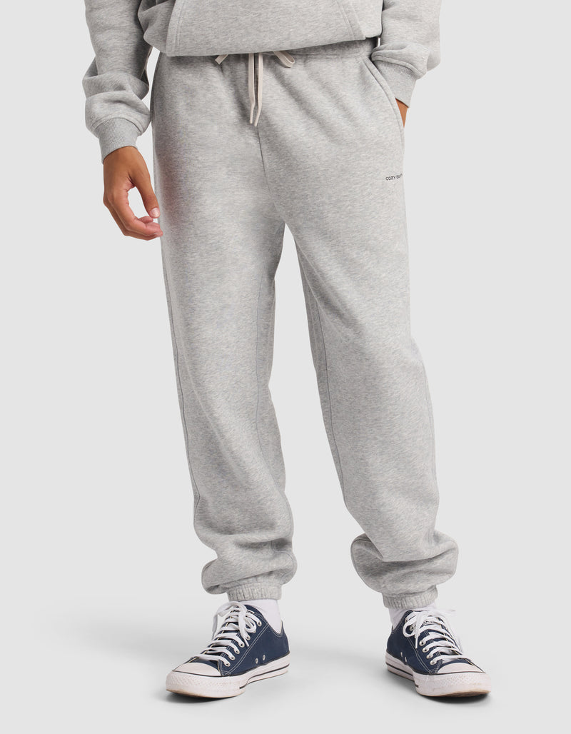 A person wears Cozy Earth Men's Cityscape 2.0 Sweatpant in light gray with a matching sweatshirt, standing against a plain background and sporting black and white Converse sneakers.