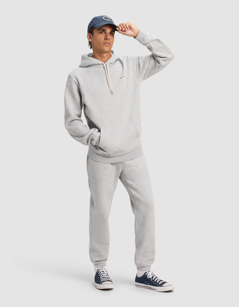 A person wearing Cozy Earth Men's Cityscape 2.0 Sweatpant in light gray, paired with a matching hoodie, dark sneakers, and a blue cap stands against a plain background, one hand in pocket and the other adjusting their cap.