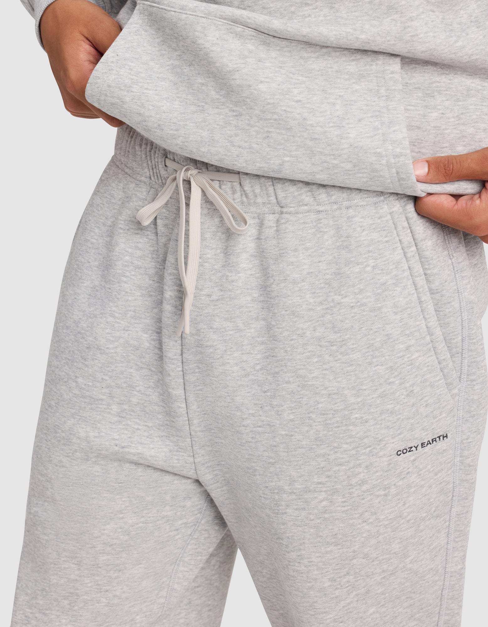 A person adjusts their sweatshirt hem while wearing Cozy Earth's Men's Cityscape 2.0 Sweatpant in light gray, featuring a drawstring waist and “COZY EARTH” text on the pocket.