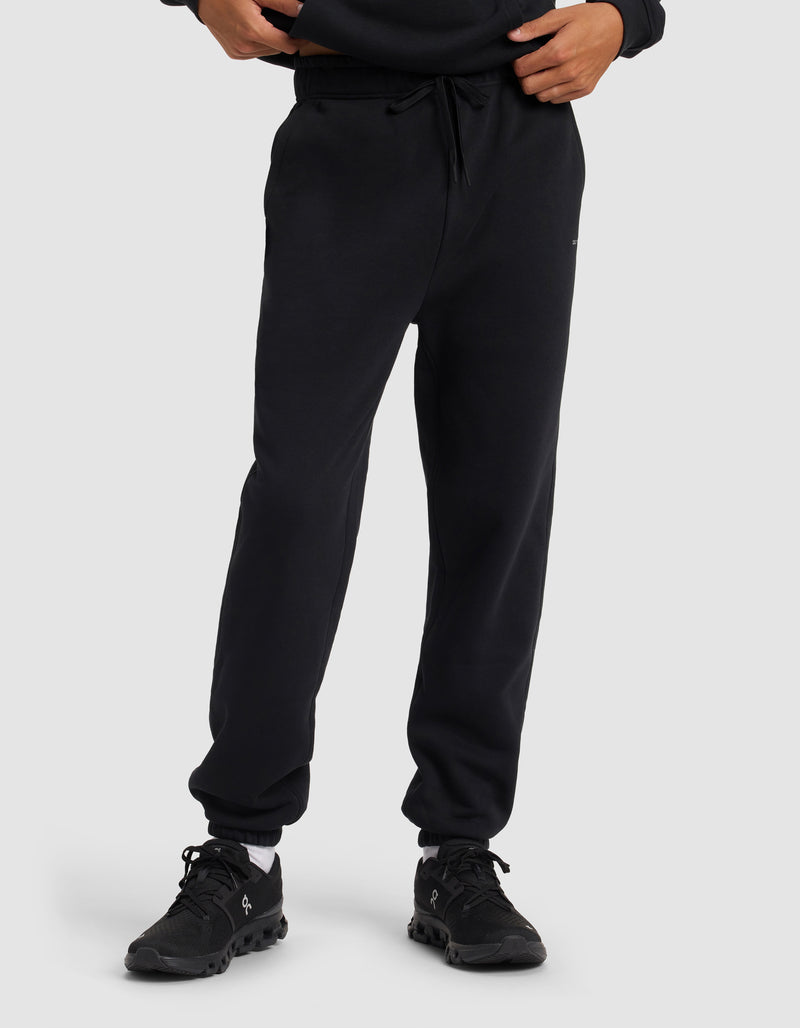 A person models the Cozy Earth Men's Cityscape 2.0 Sweatpant in black with matching sneakers, standing against a plain light gray background. Only the lower torso and legs are shown.