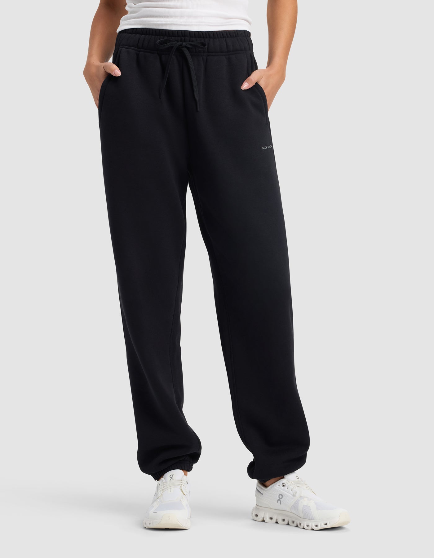 A person wearing Cozy Earth's Women’s Cityscape 2.0 Sweatpant in black, paired with a white shirt and white sneakers, stands with hands in pockets against a plain gray background.