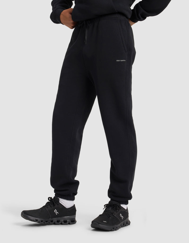 A person wears Cozy Earth Men's Cityscape 2.0 Sweatpants in black, paired with a black sweatshirt and black athletic shoes with white socks, standing against a plain light gray background. Only their lower half is shown.