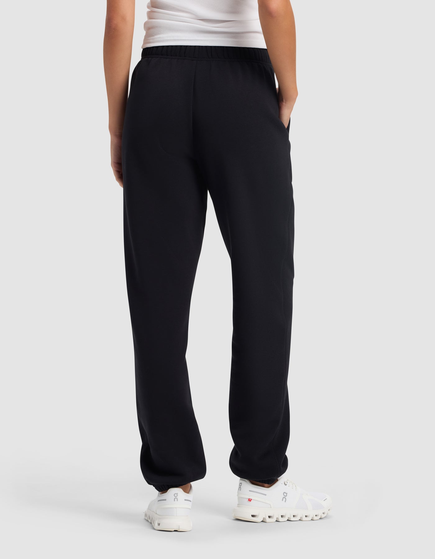 A woman wearing Cozy Earth's Women's Cityscape 2.0 Sweatpant in black, a white top, and white athletic shoes stands with her back to the camera, hands in pockets, against a plain light background.