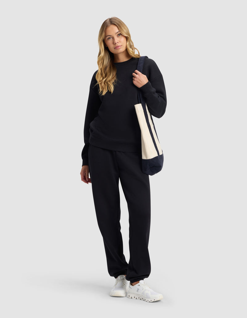 A woman with long blonde hair wears the Cozy Earth Women's Cityscape 2.0 Sweatpant in black, a matching sweatshirt, white sneakers, and carries a beige tote bag, standing against a plain light gray background.