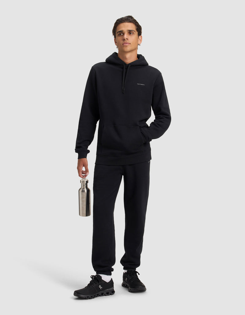 A person stands against a plain background, dressed in black with the Cozy Earth Men's Cityscape 2.0 Sweatpant and matching hoodie, black sneakers, holding a silver water bottle in their right hand and their left hand in their pocket.