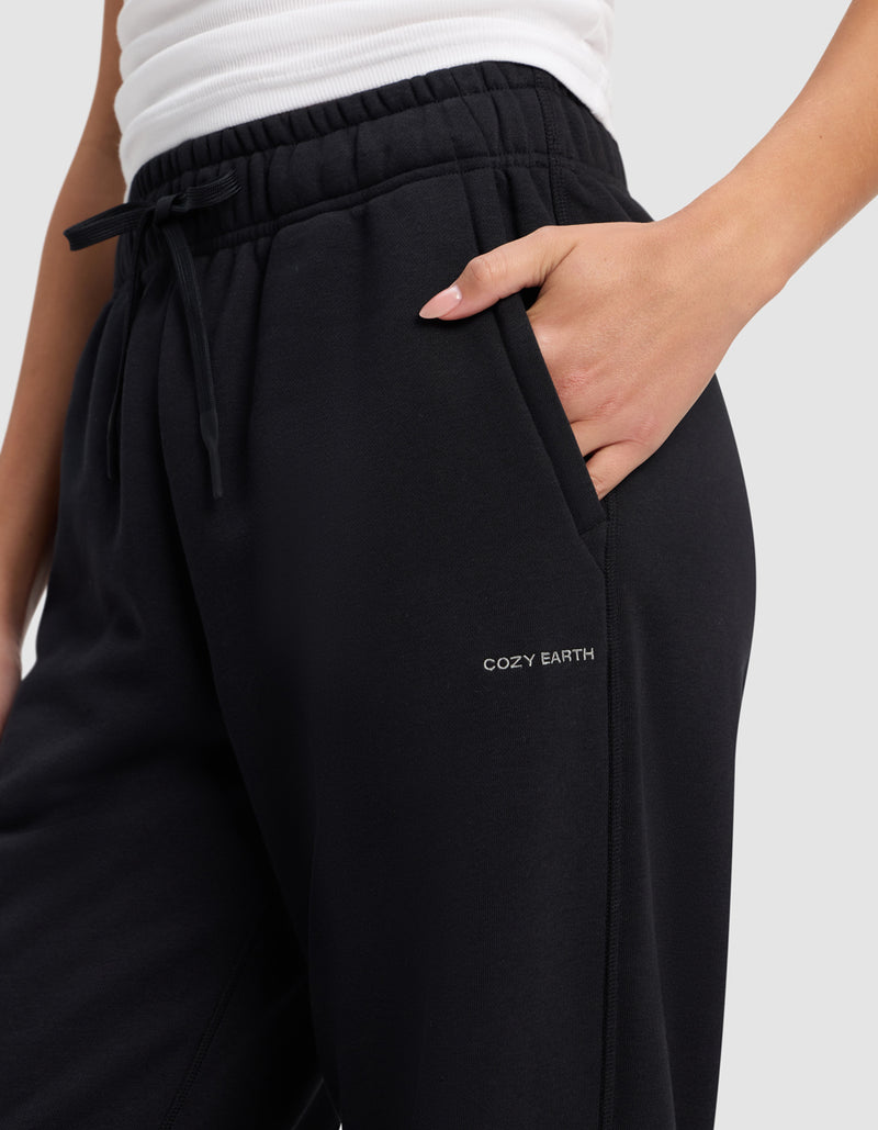 A person wearing black Cozy Earth Women's Cityscape 2.0 Sweatpants with an elastic waistband, drawstring, a white top, and one hand in the pocket. "COZY EARTH" is printed on the upper thigh of the pants.
